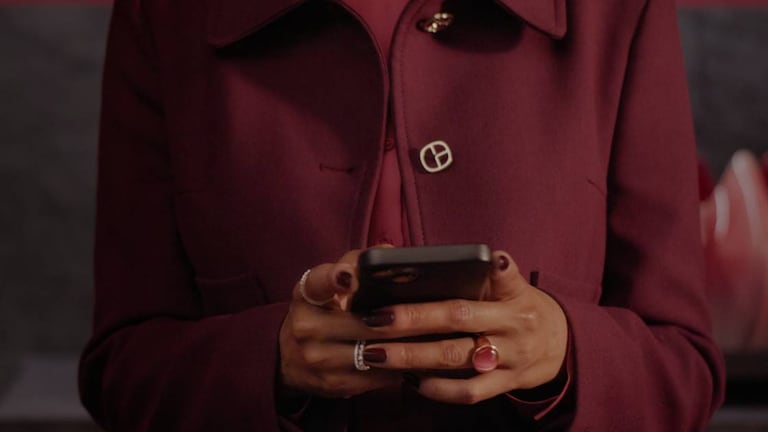 Primer plano de una persona con abrigo rojo oscuro, uñas pintadas y anillos, usando un teléfono móvil