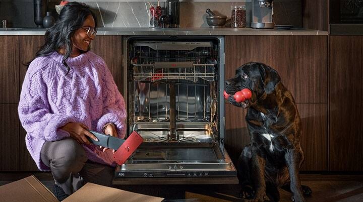 Mujer desempaquetando repuestos frente al lavavajillas, mientras sonríe a un perro negro